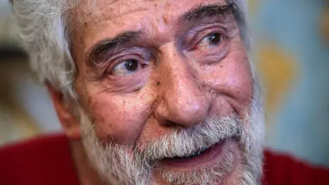 AFP Georges Abdallah, a grey-haired 74-year-old with grey moustache and beard, sits in his prison cell in a red shirt