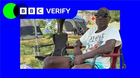 BBC A man in a white t-shirt and aqua shorts and black sunglasses sits in a camping chair on a patch of grass. A second camping chair is next to him, and tents and a wheelchair are visible in the background. In the far distance, a large building is visible.