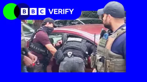 BBC A police officer leans through the smashed window of a red car to arrest a man as two others watch on.