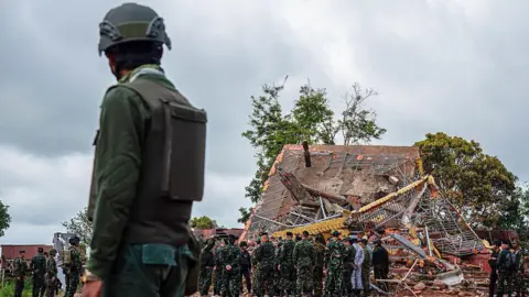 Getty Images A Cambodian soldier stands on guard near a destroyed building at the border between Thailand and Cambodia. He watches a team of soldiers and officials inspecting the site.   