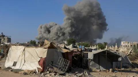 Reuters Ramshackle tent-like structures, one has a kind of a metal grid in front, with a large plume of smoke behind them and two half-finished barely-standing building on either side, in Gaza City on 24 August 2025