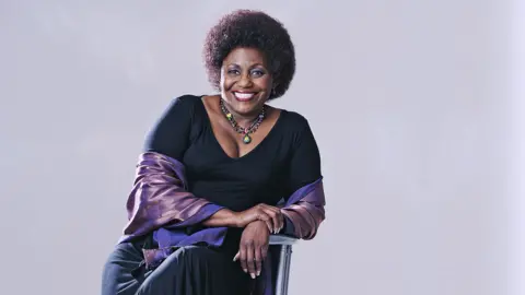Media 24 / Gallo Images A portrait style image shows actress Nandi Nyembe posing for the camera while smiling and sitting on a chair in a photography studio against a grey background, taken in South Africa in 2007.