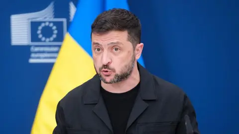 Getty Images Ukrainian President Zelensky talks to the press while standing in front of a blue and yellow Ukrainian flag. 