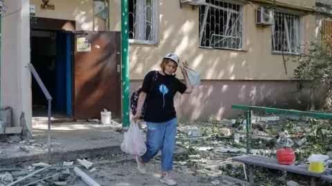 EPA A woman wearing a black top, jeans, trainers and a hard hat, and holding two plastic bags in both hands emerges out of a house after a Russian attack. Debris can be seen all around.