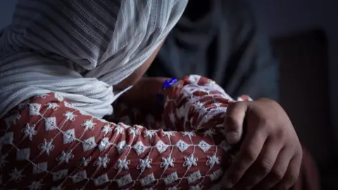 BBC Young woman wears a white headscarf with her arms crossed over her knees.