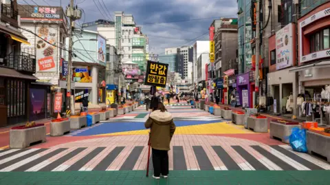 Getty Images A lively and colorful pedestrian street in Seoul, filled with vibrant crosswalk designs, unique shops, and bustling activity. A woman stands in the middle of a zebra crossing in a winter jacket carrying the sign 'escape room, half price' 