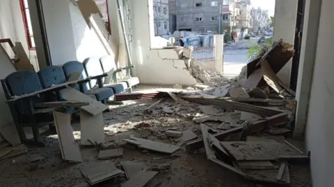 Palestine Red Crescent Society A badly damaged wall of an office, debris is scattered inside it and dust covers a bank of witting room chairs  
