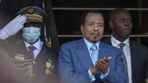 AFP via Getty Images Cameroon President Paul Biya applauds during the May 20 parade marking the 51st celebration of Unity Day in Yaounde on May 20, 2023. He is wearing a blue suit and tie. A soldier is saluting behind him.