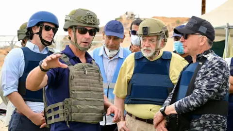 US government Mike Huckabee (second from right) and Steve Witkoff (right) stand in bullet-proof vests listening to a man wearing a helmet and sunglasses as he gestures with his hand at an aid distribution site in Gaza. 