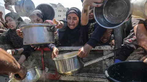MOHAMMED SABER/EPA Several people holding pans to fill with food at an aid distribution site - with one woman in the middle wearing all black visibly upset and crying