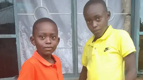 Carolyne Odour Carolyne Odour's sons - 12-year-old Daniel on the right in a yellow T-shirt and on the left, nine-year-old Elijah. They are standing outside in front of a window with a white curtain drawn.
