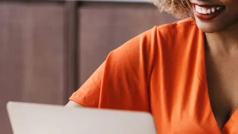 Getty Images A cropped image showing the lower half of a woman's smiling face as she looks at a laptop. She is wearing an orange top.