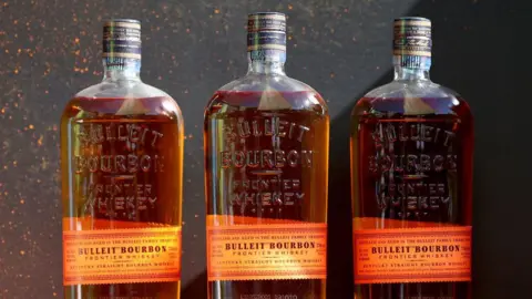Getty Images Three bottles of Bulleit Bourbon are lined up against a black stone background

The labels are orange and printed with vintage-style writing, while the bottles, filled with golden liquor, are embossed with the words "Bulleit Bourbon: Frontier Whiskey"