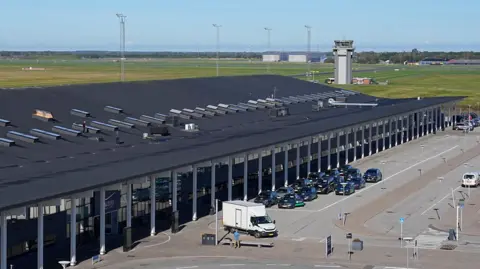 EPA An airport terminal with a black roof is pictured from above, with a series of black vehicles parked in front of the building. The airport control tower can be seen in the distance.