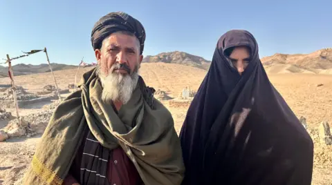Aakriti Thapar/BBC Ghulam and Nazo stand with a desert landscape behind them. He has a white beard, a turban and a green scarf wrapped around his neck. She is covering most of her face in a black shawl which is wrapped around her body