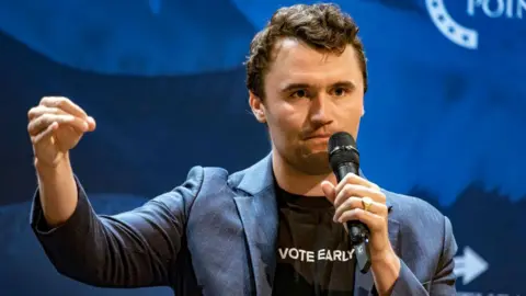 Getty Images Charlie Kirk at an event holding a microphone. His T-shirt , worn under a blazer, says 'vote early'.