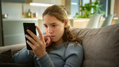 Getty Images A teenage girl sitting on a sofa looking at her phone with a worried look on her face. She is biting her fingernail.