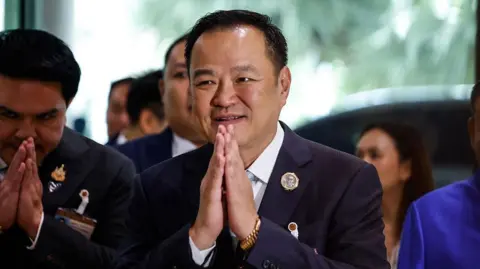 Getty Images Anutin Charnvirakul presses his palms together and greets people with a smile, while wearing a dark suit and gold watch.