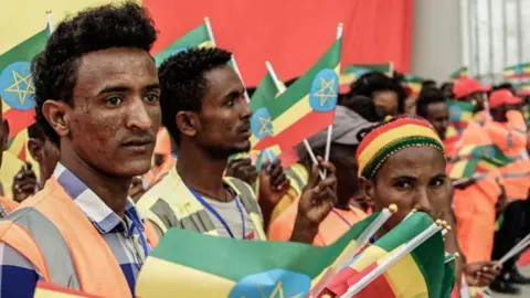 AFP via Getty Images A crowd of Ethiopian workers wearing orange hi-viz work clothes wave Ethiopian flags.