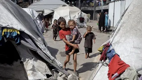 Anadolu via Getty Images A smiling older child carries a younger girl in her arms in a displaced persons camp in Khan Younis, Gaza. The pair are surrounded by dirty grey and white tents with items of clothing drying on the outside. there are other children and women in background.
