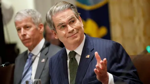 Getty Images US Treasury Secretary Scott Bessent, in a blue pinstripe suit with American flag on his lapel, raises his eyebrows and holds out his hand as he speaks during a meeting at the White House on October 14, 2025 in Washington, DC