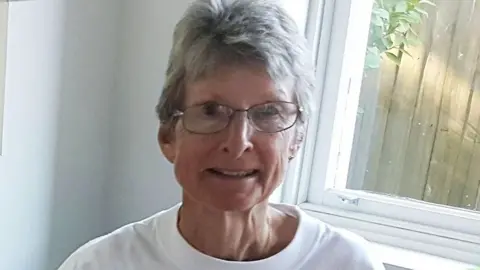 Supplied Suzanne Rees, wearing a white t-shirt and black wire glasses smiles in a photo