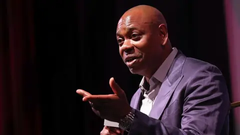 Getty Images Dave Chappelle speaks at the premiere of "Dave Chappelle: Live In Real Life"  during the 2025 Martha's Vineyard African American Film Festival at Martha's Vineyard Performing Arts Center on August 08, 2025 in Massachusetts.  He is wearing a purple suit and holding a microphone.