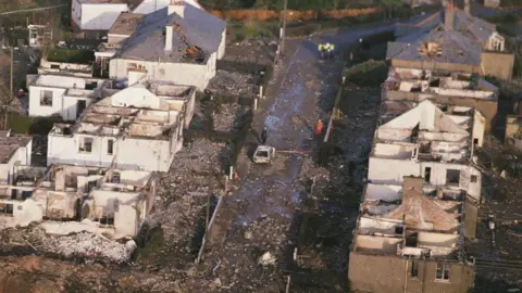 Getty Images A high view of a row of destroyed houses in Lockerbie. Their roofs have been blown off, their interiors destroyed and debris has been thrown across the land. A burnt-out car can be seen in the middle of the street and there are a few police and emergency workers just visible in the street.