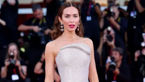 Getty Images Emily Blunt stands on a red carpet. She is wearing a silver dress with earrings.