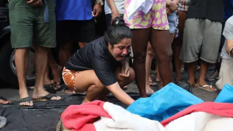 Bruno Itan A woman lifts a blue tarpaulin which covers a body. She appears to be crying out. The legs of bystanders can be seen behind her. 