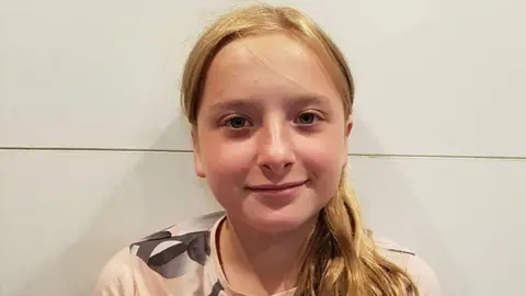 Reuters Lola Daviet is photographed standing against a white wall. She has blonde hair in a pony tail and is wearing a light pink t-shirt with grey and white detailing on the shoulder.