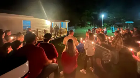 BBC A crowd holds candles and listens to a speaker outside a small building