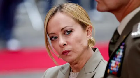 Reuters Giorgia Meloni, wearing a beige suit and gold, floral earrings, leans her head toward the camera. 
