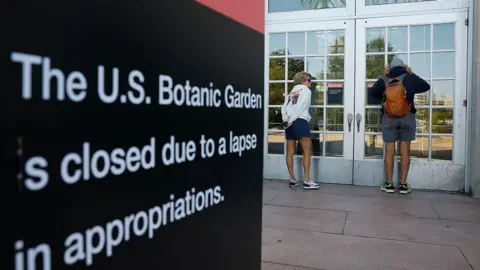 Getty Images Two visitors in shorts and sweatshirts look through the door windows of the closed US Botanical Gardens during the federal government shut down. In the foreground is a black sign with white lettering reading: 'The US Botanic Garden is closed due to a lapse in appropriations'. 