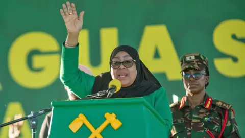 EPA/Shutterstock President Samia in a green dress and black headscarf raises her hand as she speaks at a green podium decorated by a yellow hoe and hammer, the symbol of the CCM party - behind her stands a female security officer in camouflage.