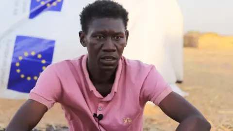 BBC Abdu-Rabbu Ahmed, wearing a short-sleeved pink shirt, with a microphone attached to it, sits in front of a white tent in Tawila that has EU logos on it.