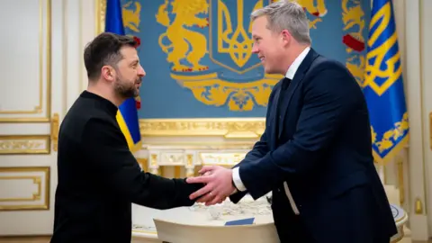 PA Media Volodymyr Zelensky, in black mockneck, puts hand forward, which Dan Driscoll, which grey hair and navy suit, grips in both hands. In the background are gold and blue insignia and flags