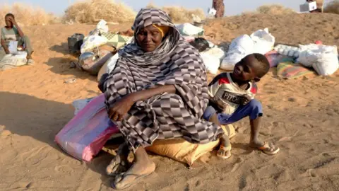 Reuters A woman wrapped in a cloth and wearing sandals looks down as she sits on a bag of her belongings, which is on sand. A small child is next to her and looks at her smiling.