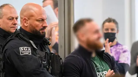 MATTHIAS RIETSCHEL/POOL/AFP Defendant Johann G (blurred and on the right)  is brought to the courtroom at the start of a trial against seven left extremists accused for membership in a criminal organization on November 25, 2025 at the Higher Regional Court of Dresden, eastern Germany