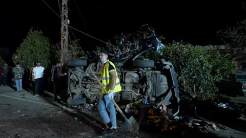 Neha Sharma/BBC A young man in hi-vis holds a shovel in front of a car, on its side and destroyed by an apparent explosion. Other young men look on. It's dark and the car is on a street in front of a hedge.