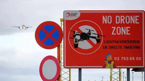 Getty Images Photo shows a sign that reads 'No Drone Zone' near the Brussels-National Airport. A Brussels Airlines passenger jet can be seen coming in to land in the background.