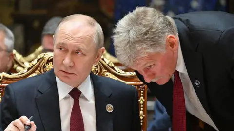 GRIGORY SYSOYEV/POOL/AFP via Getty Images Russian President Vladimir Putin in a suit and red tie say behind a table, Peskov, a man with grey hair wearing a suit is bending down listening to him talk.