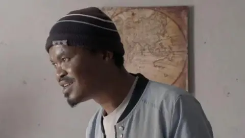 BBC A side view of the head and shoulders of Sinqhiwe Goodman Sithole. He is wearing a dark beanie hat and grey jacket.