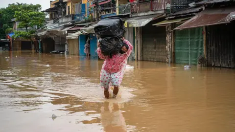 Getty Images A man in a raincoat wades through the floodwaters in Hoi An, carrying two large packs of items wrapped in black trash bags