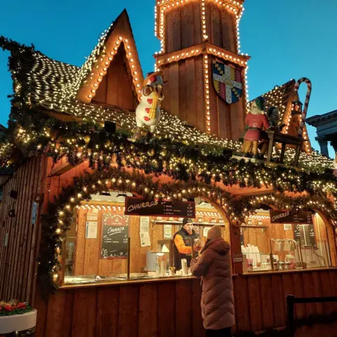 BBC A stall selling churros at a Christmas market. The wooden stall is adorned with fake Christmas tree sprigs, golden fairylights, signs saying "churros". A man stands behind the counter at the stall, serving a woman in a long winter coat