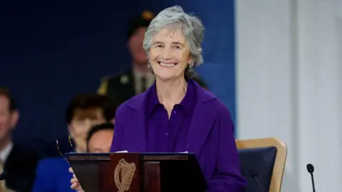PA Media Catherine Connolly, with short grey hair, wearing a purple top and purple jacket. She is standing behind a wooden podium. 