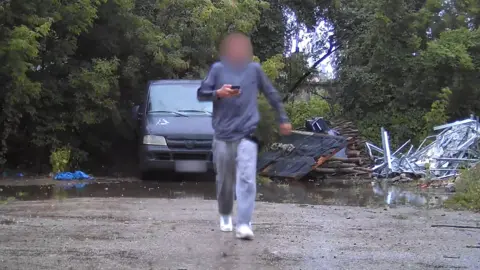 SBU A surveillance photo of a male teenager wearing grey sweat pants and shirt and with his face blurred, carrying a mobile phone. He is walking away from a grey van parked on hard surface in a wooded area. 