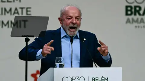 Pablo Porciuncula/AFP Brazilian President Lula stands behind a white podium with the words "Belém COP30". On the podium is a glass of water. Lula is in a blue suit his hands pointing forward and his mouth open mid-speech.
