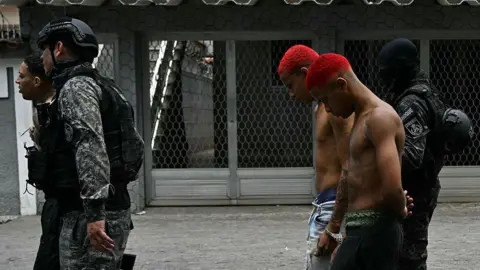 MAURO PIMENTEL/AFP via Getty Images Police officers escort suspects arrested during the Operacao Contencao (Operation Containment) out of the Vila Cruzeiro favela, in the Penha complex, in Rio de Janeiro, Brazil, on October 28, 2025. 