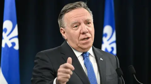 Bloomberg via Getty Images Quebec Premier Francois Legault, wearing a dark suit and a blue and white patterned tie, speaks into two microphones. Behind him, against a black backdrop, are two blue and white Quebec flags. 